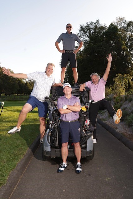 four men postioned around and on a golf cart. One is standing on the top of the cart with his hands on his hips, another wearing a cap is standing in front of the golf cart with folded arms, and two are on either side of the cart with only one foot on it and the other foot in the air
